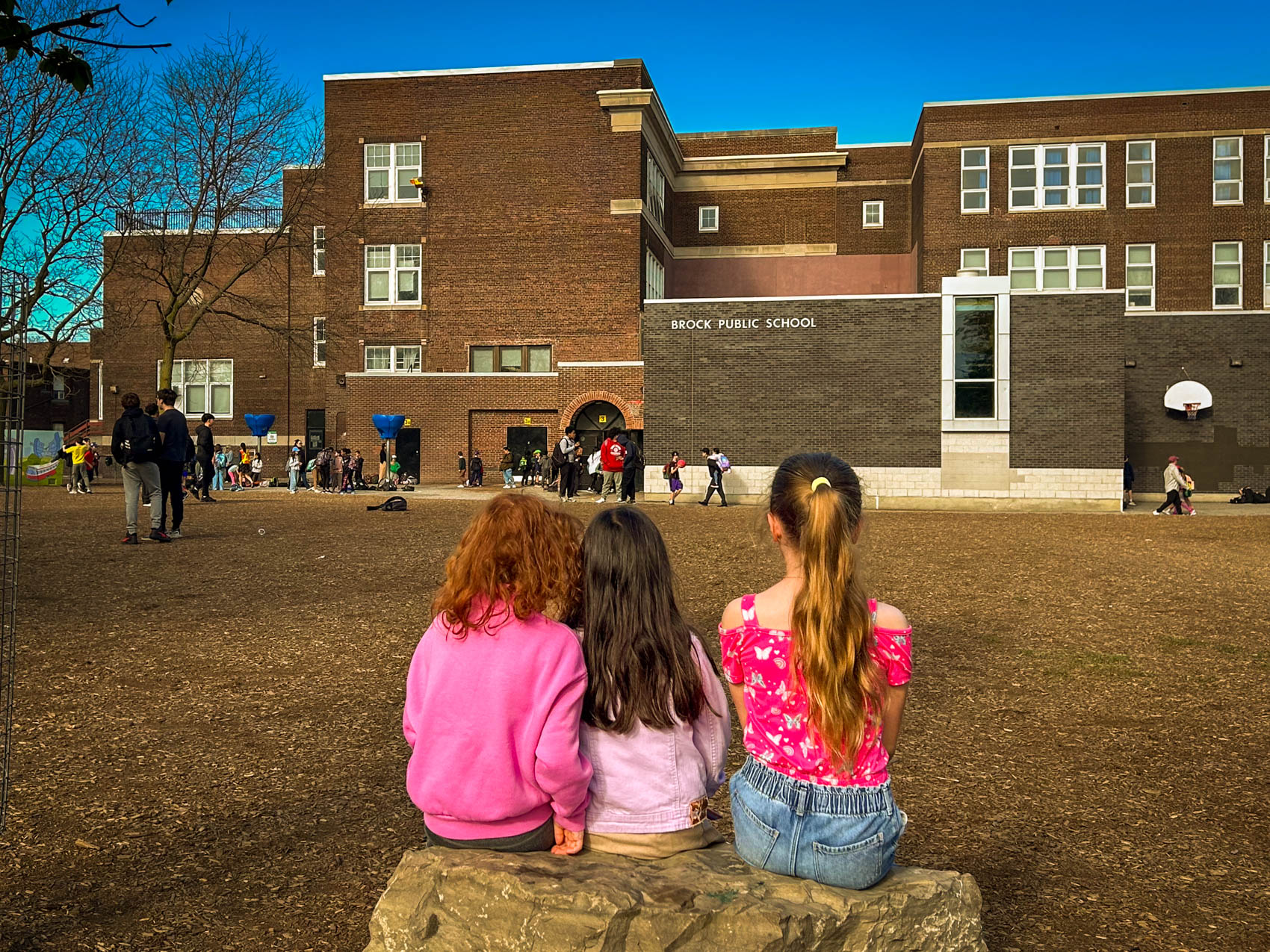 Three friends sitting on a rock in front of the Brock Yard, with the school in the background