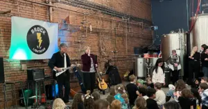 Photo of Lori and Kurt performing, children sitting and watching, Brock School banner on wall
